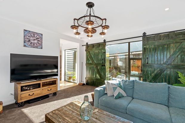 Bright living room with a large blue sectional sofa, rustic timber coffee table and media console, wall-mounted television, and distinctive weathered green barn-door shutters covering wide glazing. A decorative wagon-wheel chandelier and white walls create a relaxed rustic-coastal character.