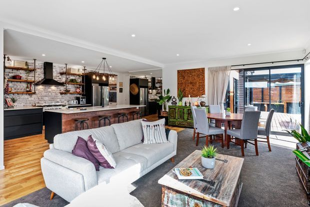 Open-plan contemporary living space combining a kitchen, dining area and lounge. The kitchen features dark cabinetry, a white brick-look splashback, stainless-steel appliances, a central island and pendant lighting. A light upholstered sofa faces a rustic timber coffee table, while the dining area opens through large glazed doors to an outdoor deck. Decorative timber artwork and a green sideboard add warmth and character.