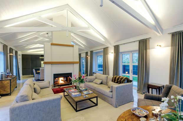 Bright, spacious open-plan living room with a high vaulted ceiling and exposed white-painted structural beams. The room features a central fireplace with a simple built-in surround, two large upholstered sofas, coffee table, armchair, side tables, floor-to-ceiling curtains, and extensive glazing. Neutral grey, cream, and white finishes create an airy, refined atmosphere.