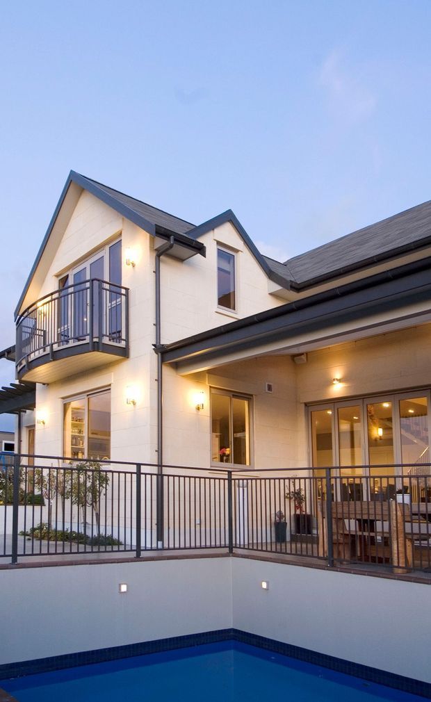 Exterior view of a two-storey contemporary home at dusk, featuring a light rendered facade, dark pitched gabled roofs, a curved metal balcony, extensive outdoor railings, warm exterior lighting, and a partially visible swimming pool in the foreground.