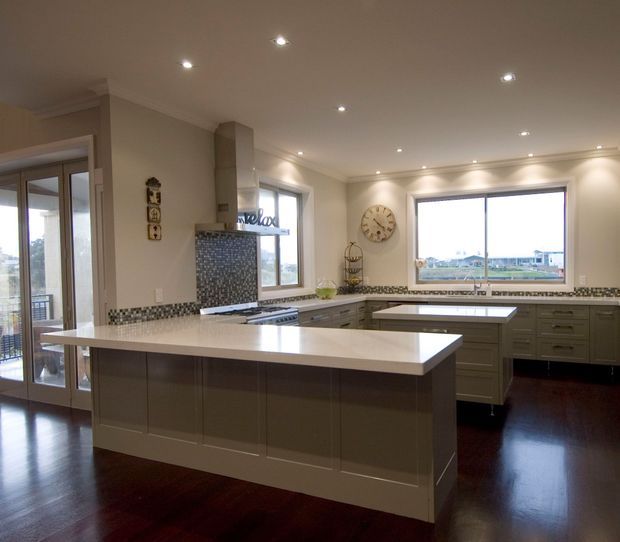 Spacious traditional kitchen with a large central island, dark timber flooring, grey-green shaker-style cabinetry, pale stone benchtops, mosaic tile splashback, stainless steel appliances and extensive windows and glazed doors. Recessed ceiling lighting and a neutral palette create a bright, refined family kitchen.