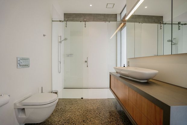 Contemporary bathroom featuring a wall-mounted toilet, frameless glass shower enclosure with exposed sliding hardware, a vessel basin on a long dark benchtop, mirrored wall cabinets, and warm timber vanity cabinetry. The floor is finished in a speckled terrazzo-like surface, with predominantly white walls and restrained modern detailing.