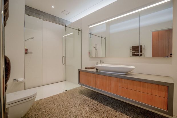 Contemporary bathroom with a large frameless glass shower enclosure, wall-hung toilet, floating timber-toned vanity with a dark benchtop, and a vessel basin. A full-width mirror and integrated linear lighting create a clean, spacious appearance. The floor is finished in a speckled neutral surface, while warm wood cabinetry adds contrast to the predominantly white and grey palette.