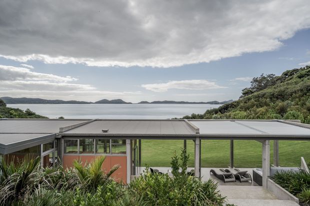 Exterior view of a low-profile modern house overlooking a calm bay or coastal inlet. The image shows a broad flat roof, extensive glazing, concrete or grey structural elements, a lawn and outdoor seating terrace, with dense landscaped planting in the foreground and a sloping vegetated site to the side. The distant water and islands are visible beyond the property under an overcast sky.