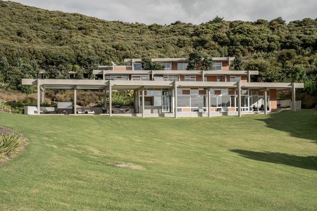 Wide exterior view of a long, low-slung modern house set against a densely vegetated hillside. The residence features layered flat roof forms, extensive horizontal glazing, pale rendered walls, warm timber-toned panels, and a large open pergola or covered outdoor living structure facing a broad lawn.