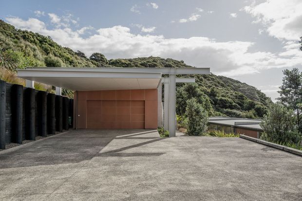 Exterior view of a modern flat-roofed structure on a steep, heavily vegetated hillside. A broad concrete-paved forecourt leads to a covered carport or entry canopy, with a warm terracotta-toned wall panel and slender white structural posts. Dark vertical retaining elements line the left side, while additional low roof forms and dense greenery extend down the slope.