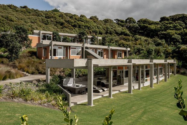 Exterior view of a contemporary multi-level hillside residence integrated into dense native vegetation. The house features broad horizontal concrete slab roofs and terraces, slender concrete columns, extensive glazing, warm timber-toned wall panels, and a large manicured lawn. Outdoor seating and a spa are visible beneath the covered terrace.