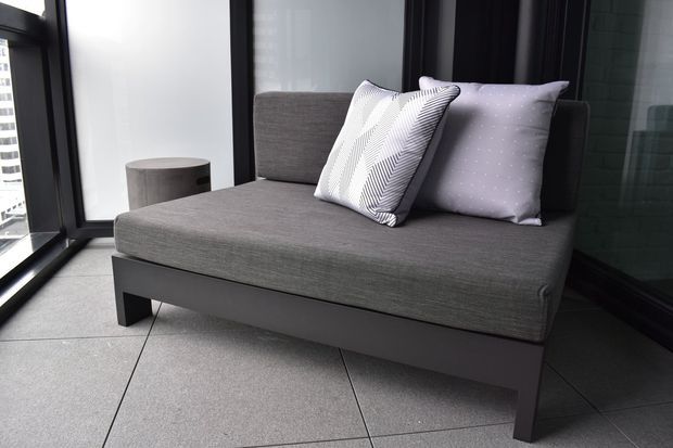 A compact contemporary seating nook featuring a charcoal-grey upholstered sofa with a dark timber or metal base, patterned cushions, and a small round side table. Floor-to-ceiling black-framed glazing surrounds the space, with large light-grey tiled flooring and a dark reflective wall surface creating a refined, urban feel.