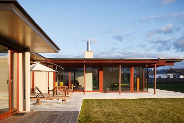 Single-storey contemporary residential courtyard exterior with broad flat roof planes, extensive floor-to-ceiling glazing, warm timber-toned window and door frames, and a paved outdoor terrace furnished with dining and lounge seating. A small lawn occupies the foreground, with the low horizontal house opening toward the outdoor space.