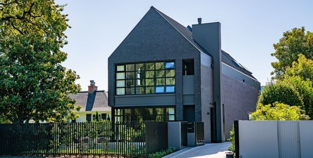 Exterior view of a contemporary multi-storey residence with a steep gabled roof, dark brick facade, large black-framed grid windows, and a tall central chimney-like vertical volume. A dark metal fence and driveway frame the frontage, with mature trees and neighbouring homes visible around the property.