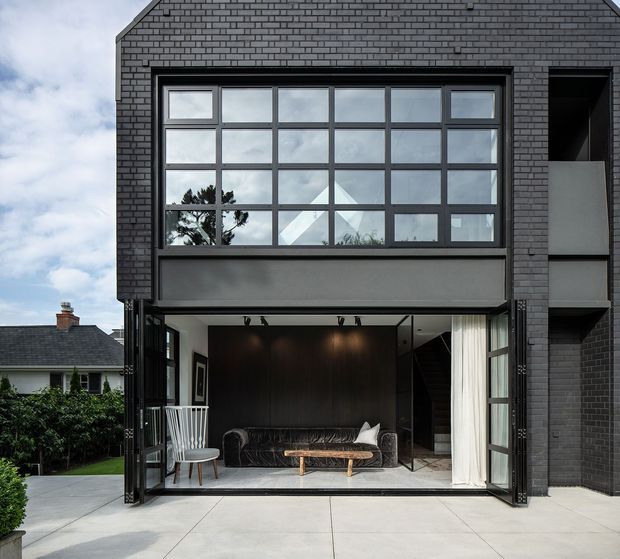 A contemporary two-storey dark brick residence viewed from the rear courtyard. The facade features a large multi-pane upper-level window, a wide fully glazed lower-level opening with bi-fold or folding doors, and a covered outdoor living connection. A furnished living room with a dark sofa, timber coffee table, dining chair, dark feature wall and staircase is visible through the open glazing. The courtyard has pale concrete paving and the image also shows part of an adjacent traditional house.