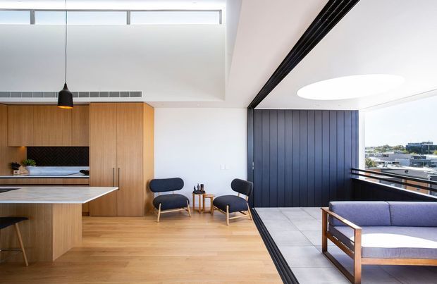 Bright contemporary open-plan interior combining a kitchen, dining or living area, and an enclosed balcony or terrace. The space features light timber flooring and cabinetry, a large island with a pale stone benchtop, black pendant lighting, a dark vertically panelled feature wall, expansive glazing, and a circular skylight or ceiling opening. Mid-century-inspired lounge chairs and a timber-framed sofa occupy the living area, with an urban outlook beyond the glazing.