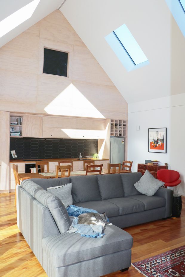 Bright open-plan living and dining area with a compact kitchen at the rear. The room features a steeply vaulted ceiling with skylights, a large pale timber-lined triangular wall, extensive light timber cabinetry, a dark hexagonal tile splashback, polished timber flooring, a large grey sectional sofa, a timber dining table, and colourful contemporary furnishings and artwork.