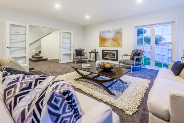 Bright, spacious living room with white walls and trim, plush grey carpeting, a large cream shag rug, cream upholstered sofas, patterned accent chairs, and a dark sculptural coffee table. A wide glazed door opens to an outdoor balcony or terrace, while built-in shelving and a staircase are visible at the rear. Contemporary artwork, recessed ceiling lights, and geometric textiles add visual interest.