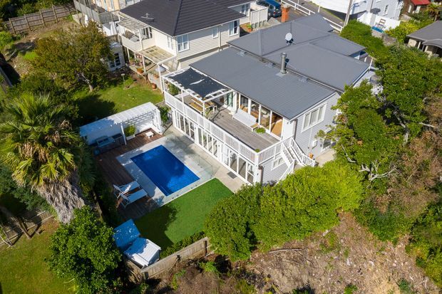 Aerial view of a multi-level residential property on a landscaped, sloping site. The home has grey pitched roofing, light-coloured weatherboard-style cladding, extensive white-framed glazing, multiple timber decks and balconies, and an outdoor swimming pool with surrounding paved areas and lawn. Adjacent homes are visible in the background.