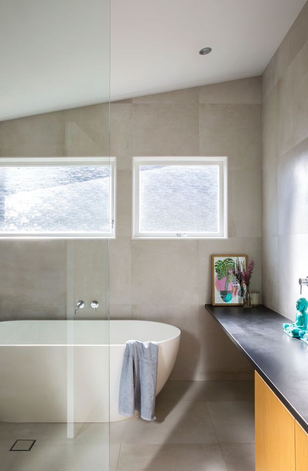 Bright contemporary bathroom featuring a freestanding white oval bathtub behind a frameless glass shower screen, large beige stone-look wall and floor tiles, and a dark countertop with warm timber vanity cabinetry. Two frosted windows provide natural light, while a small framed artwork and decorative objects add colour.