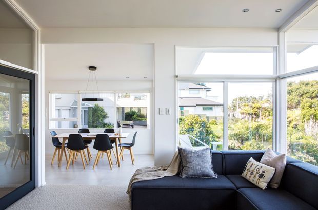Bright contemporary open-plan living room with a dark sectional sofa in the foreground, opening directly to a dining area with a round table and chairs. Large windows and glazed doors provide abundant daylight and views to a leafy suburban setting. The interior has white walls and ceilings, light carpeting, simple recessed lighting, and dark-framed glazing.