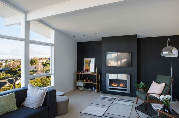 A contemporary living room with large corner windows overlooking a suburban landscape. The room features a dark charcoal fireplace wall with a built-in electric or gas fire, a neutral sofa and armchair, a geometric area rug, a sculptural floor lamp, and simple open shelving. The palette is restrained in charcoal, white, grey, and muted green, with abundant daylight and clean-lined furnishings.