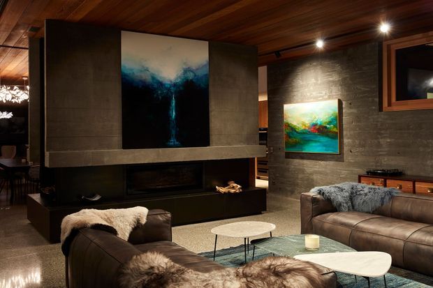 A moody, sophisticated living room centred on a large concrete fireplace feature wall with integrated firebox and broad mantel. Dark leather sofas, fur throws, low sculptural coffee tables, timber ceiling boards, recessed spotlights, artwork, and timber-accented cabinetry create a warm contemporary lodge-like atmosphere. A dining area is partially visible beyond the living space.
