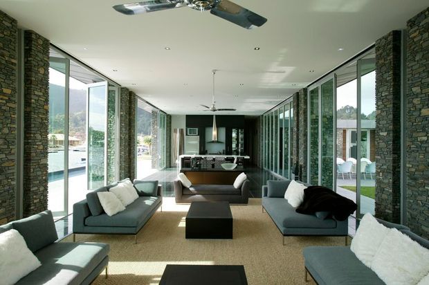 Long, bright contemporary living room with floor-to-ceiling glazed walls, exposed stacked-stone columns, pale ceiling, and multiple ceiling fans. Grey sofas with white cushions are arranged around dark rectangular coffee tables on a neutral carpet, with a compact kitchen or bar area visible at the far end. The glazing opens toward landscaped outdoor areas and a mountainous backdrop.