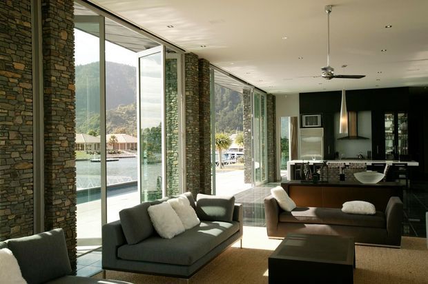 A spacious contemporary living room with full-height glazed sliding doors opening toward a pool and mountain landscape. Exposed stacked-stone structural columns frame the glazing, while dark upholstered sofas, a low coffee table, recessed ceiling lighting, and a ceiling fan create a refined indoor-outdoor setting. A dark kitchen or dining area is visible at the rear.