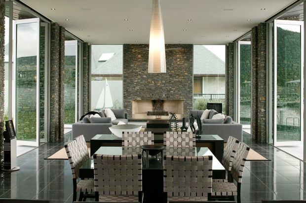 A refined open-plan living and dining room with full-height glazing and multiple open glass doors on both sides. The room features a dark polished tiled floor, exposed stacked-stone walls, a central stone fireplace, a long dark dining table with woven upholstered chairs, and two grey sofas arranged around the fireplace. A large sculptural pendant hangs above the dining area, creating a balanced, symmetrical composition with views into the surrounding landscape and adjacent buildings.