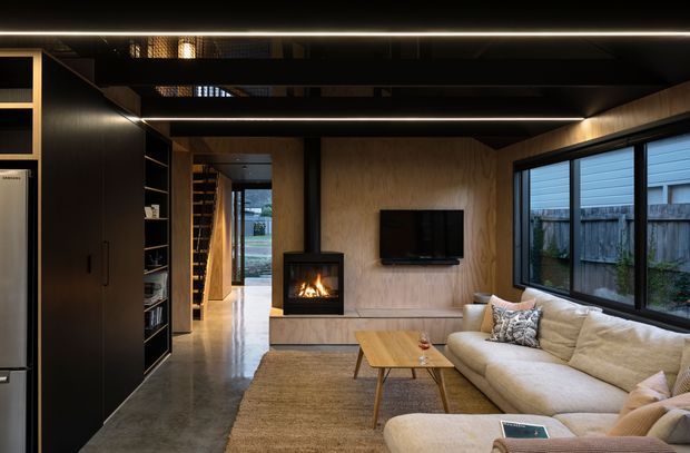 A dark, refined contemporary living room with a large cream sectional sofa, timber coffee table, wall-mounted television, and a built-in fireplace set into a warm plywood feature wall. Black built-in cabinetry lines the left side, while expansive black-framed windows provide views to a fenced exterior. Linear recessed ceiling lighting and polished concrete flooring contribute to the modern, moody interior.