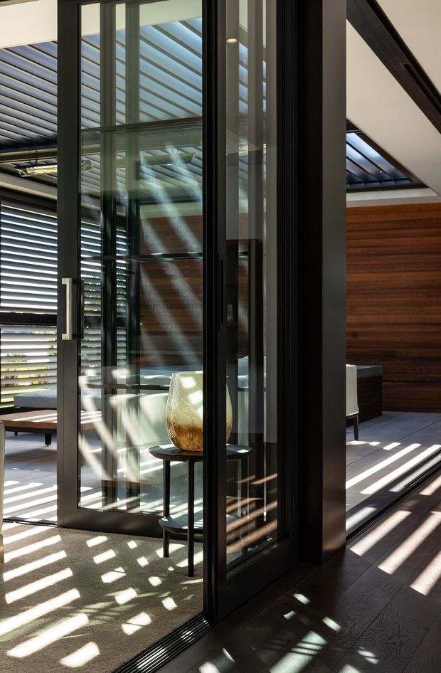 Interior view centred on a tall black-framed glazed doorway or partition, with strong sunlight casting striped shadows across dark timber flooring and a light carpeted area. A warm horizontal timber feature wall, louvred shading, clerestory-style glazing and a small side table with a decorative vessel are visible. The space has a refined contemporary architectural character.
