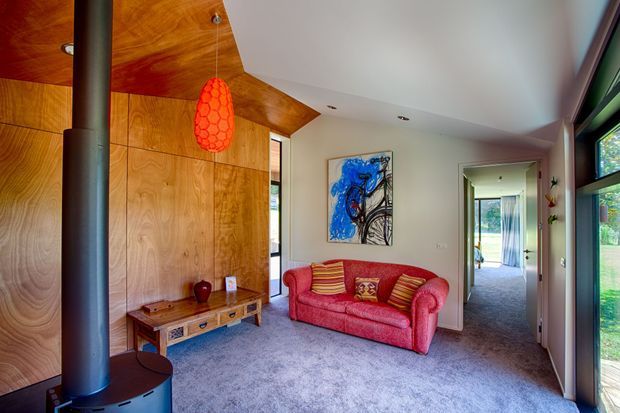 A warm, compact living room with a red upholstered sofa, grey carpet, timber-lined feature wall and ceiling, a black cylindrical wood-burning stove, and a bright orange pendant light. Abstract artwork and a timber media console add colour and texture. A hallway and glazed exterior door or window are visible to the right.