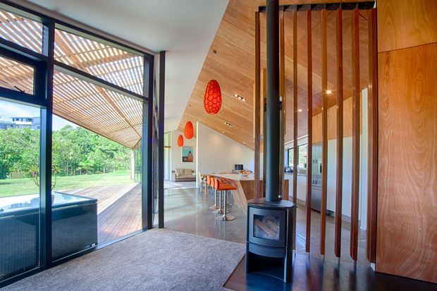 A bright contemporary open-plan living area viewed from an entry zone, with a black freestanding wood-burning stove beside a timber slat screen. The space opens through large dark-framed glazed doors to a timber deck and pool area. Inside, a warm timber-lined ceiling, polished concrete floor, kitchen island with orange bar stools, and suspended orange pendant lights create a modern indoor-outdoor setting.