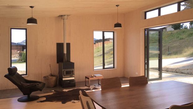 Open-plan timber-lined interior combining a living and dining area. Warm plywood walls and ceiling surround a large wooden dining table, black wood-burning stove, sculptural lounge chair, and expansive glazed doors opening to the outdoors. The room is bright and simply furnished, with a contemporary rustic character.