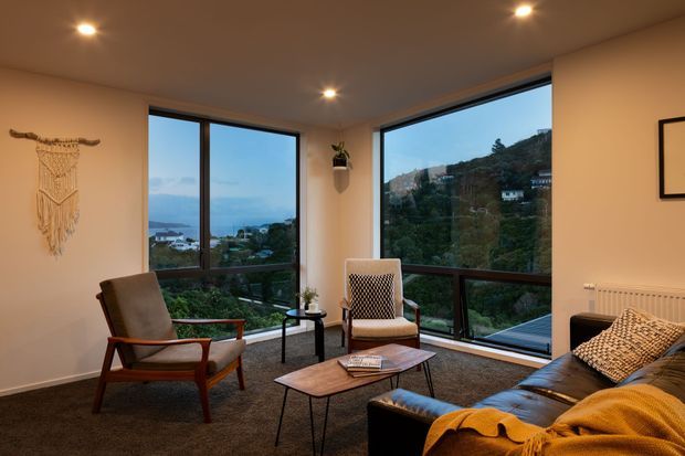 A warm, inviting living room with mid-century inspired timber-framed lounge chairs, a dark leather sofa, patterned cushions, a geometric coffee table, and a mustard throw. Large corner windows provide expansive views over a green hillside and distant water. Recessed ceiling lights, cream walls, dark carpet, and simple wall decor complete the space.