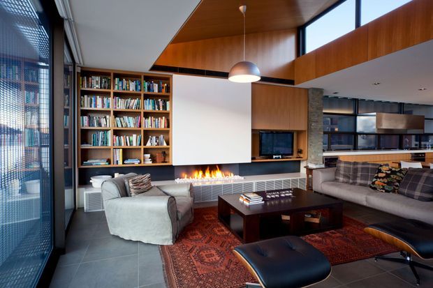 A sophisticated contemporary living room with a built-in timber library wall, linear fireplace, television niche, large sectional sofa, armchair, dark coffee table, and patterned rug. Floor-to-ceiling glazing with external screening brings in abundant daylight, while warm timber wall and ceiling panels add contrast to the dark tiled floor and black-framed kitchen glazing beyond.