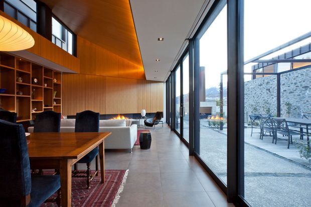 A sophisticated open-plan interior combining a dining area, living room and floor-to-ceiling glazed outdoor connection. Warm timber wall and ceiling panelling, built-in shelving, a fireplace, dark-framed glazing, stone-look flooring and contemporary furnishings create a refined, inviting atmosphere. A paved outdoor terrace and stone wall are visible beyond the glazing.
