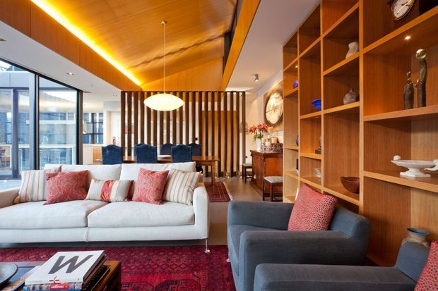 A sophisticated open-plan living and dining area with a large white sectional sofa, blue upholstered armchairs, patterned red rug and cushions, extensive timber built-in shelving, and a timber-lined ceiling. A vertical timber-slat screen separates the living zone from the dining area, while full-height glazing brings in abundant natural light. The space has a warm, richly layered, mid-century-influenced interior with curated decorative objects and artwork.