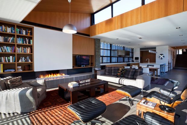 A spacious open-plan mid-century-inspired living room with extensive built-in timber bookcases and cabinetry, a linear open fireplace beneath a large white feature wall, leather lounge chairs and ottomans, a sectional sofa, and a dark timber coffee table. High clerestory glazing and a partial double-height volume bring strong natural light into the room, while the space opens toward a dining and kitchen area.