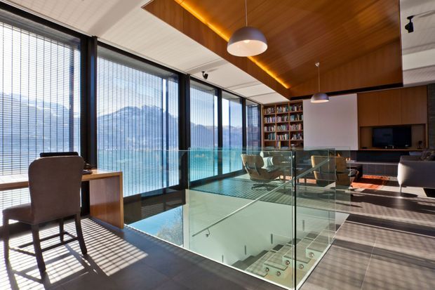 A sophisticated double-height living and study area with floor-to-ceiling glazing overlooking a mountainous landscape. The space features a glass balustrade around a stair void, a compact desk, dark upholstered seating, built-in bookshelves, a media wall, warm timber ceiling panelling with concealed strip lighting, pendant lights, and dark tiled flooring.