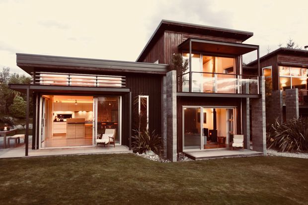 Exterior view of a contemporary multi-level residential house with dark timber cladding, concrete masonry columns, broad roof overhangs, and extensive glazed sliding doors. Warm interior lighting is visible through the glazing, with a balcony and landscaped lawn in the foreground.