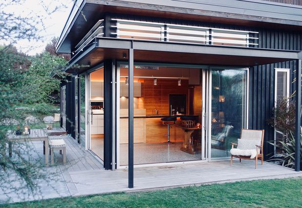 Exterior view of a contemporary dark-clad multi-level house opening onto a timber deck. Large sliding glass doors reveal a warm timber-lined interior with a kitchen or bar area, while an upper balcony has horizontal metal railings. The scene includes a paved outdoor seating area, lawn, and surrounding planting.