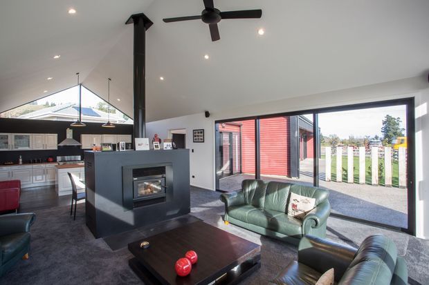 Open-plan contemporary living room with a substantial dark freestanding fireplace dividing the lounge from the kitchen and dining area. The room has a white ceiling with recessed downlights and a ceiling fan, dark carpet, large glazed sliding doors opening to an outdoor area, and green leather seating around a dark coffee table. A white kitchen with pendant lighting is visible in the background.