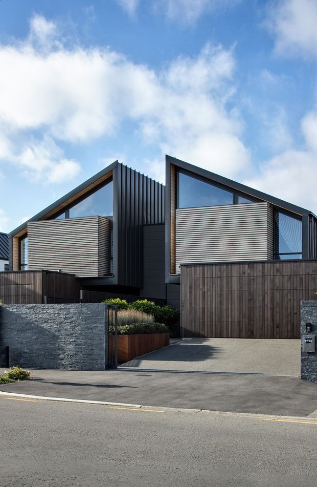 Street-facing exterior view of a contemporary multi-level residential building with two angular gabled roof forms, dark vertical metal cladding, horizontal light-toned timber or fibre-cement wall panels, large glazed openings, and tall timber boundary fencing. A paved driveway, dark stone masonry walls, and low planting define the frontage.