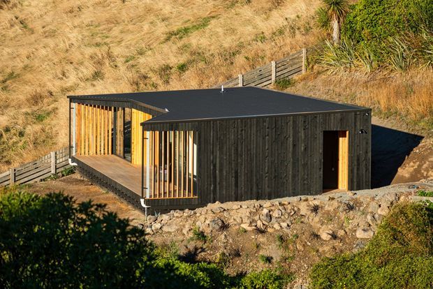 A compact, single-storey contemporary building set into a sloping, dry grass-covered site. The structure has dark vertical timber cladding, a low flat roof, warm timber slatted screening around glazed openings, and a simple timber entry door. Stone edging and retaining fencing define the immediate landscape.