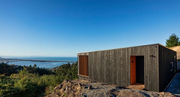 A compact contemporary flat-roofed cabin clad in dark vertical timber boards, positioned on a rocky elevated site with expansive coastal and water views. The simple rectangular form includes a recessed timber entry and narrow side glazing, surrounded by native vegetation under a clear blue sky.