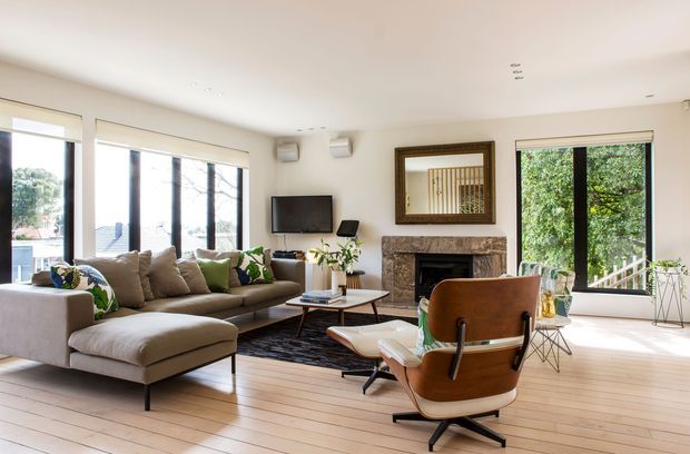 Bright, spacious living room with large windows, pale timber flooring, a light sectional sofa, mid-century style lounge chair, low coffee table, wall-mounted television, and a stone-clad fireplace. The room opens visually to leafy outdoor surroundings and uses a warm neutral palette with abundant natural light.
