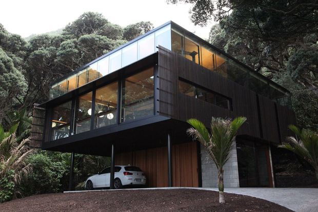 A contemporary elevated home set within dense native bush, featuring a dark timber-clad upper volume, extensive floor-to-ceiling glazing, slender structural supports, and a partially enclosed carport beneath. Warm interior lighting is visible through the windows, with a gravel driveway and tropical planting in the foreground.