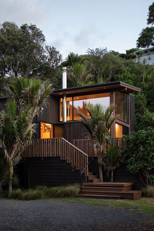 Exterior view of a compact, elevated dark timber-clad dwelling nestled among dense coastal vegetation. The house has a low-pitched roof with broad eaves, extensive floor-to-ceiling glazing, warm interior lighting, an upper-level terrace or deck, and timber stairs and balustrades leading up from a gravel driveway. Palm-like native planting surrounds the structure, creating a secluded bush setting.