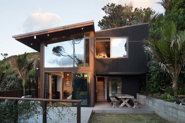 Exterior view of a compact two-storey contemporary house set into dense landscaped vegetation. The dark-clad upper volume contrasts with warm timber framing and large floor-to-ceiling glazing, while a broad mono-pitched roof projects over the living area. A timber deck, outdoor dining setting, concrete retaining wall and sloped garden surround the facade.