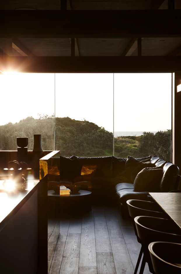 Dark, atmospheric open-plan living and dining space with a large black sectional sofa facing expansive floor-to-ceiling glazing. The room features dark timber flooring, a long dining or kitchen counter with integrated seating, exposed timber ceiling structure, and sculptural ceramic objects silhouetted against a sunlit coastal or vegetated view.