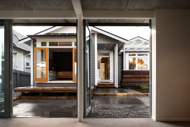 View from inside a modern dwelling through large glazed openings toward a covered outdoor courtyard and several connected small gabled structures. The architecture features grey weatherboard cladding, dark-framed glazing, timber decking, concrete paving, gravel, and a warm timber-framed entry door. A textured exposed concrete ceiling and white structural beams are visible inside.