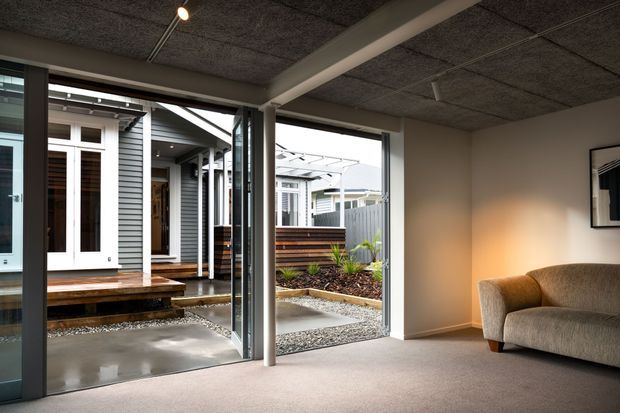 A contemporary living room with neutral carpet, a textured dark ceiling, exposed structural beams, and a curved upholstered sofa. Large sliding glass doors open directly onto a sheltered courtyard with concrete paving, gravel, timber screening, and a covered outdoor connection to the adjoining building.
